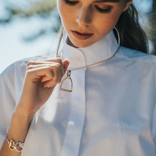 Equestrian woman wearing a gold necklace with a horse stirrup pendant, outdoors.